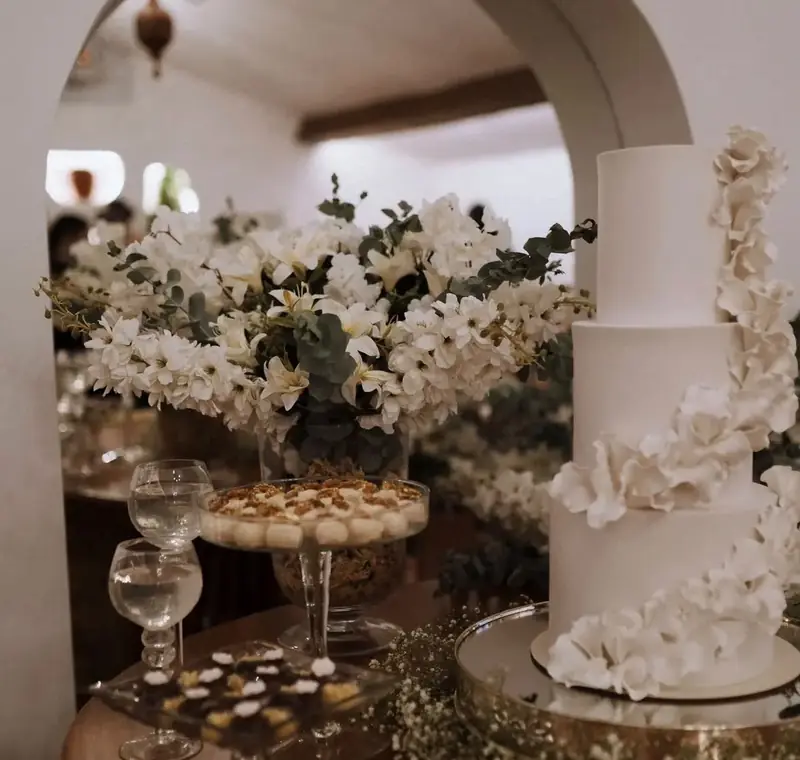 Bolo de casamento branco com flores de açúcar, taças e arranjo de flores brancas e eucalipto