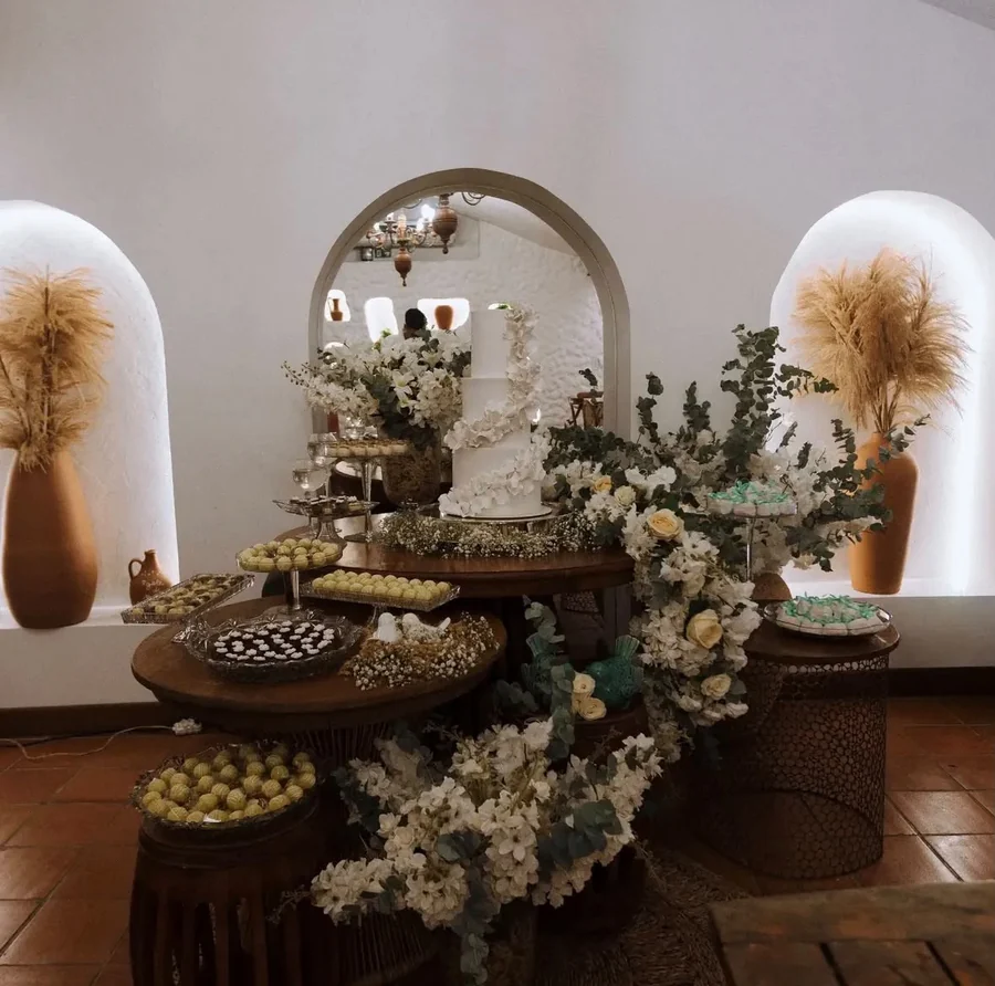 Mesa de doces montada no Mezanino, com bolo, arranjos de flores e os nichos acesos ao fundo